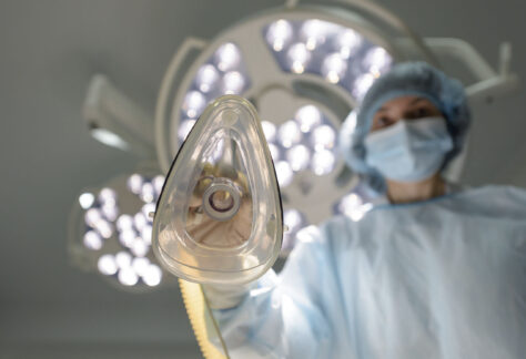 Blickwinkel aus der Perspektive eines Patienten auf dem OP-Tisch: Eine medizinische Fachkraft in blauer OP-Kleidung, Haube und Mundschutz hält eine Anästhesiemaske über die Kamera. Im Hintergrund sind helle Operationsleuchten zu sehen. Die Szene vermittelt den Moment vor der Narkoseeinleitung in einem Operationssaal.