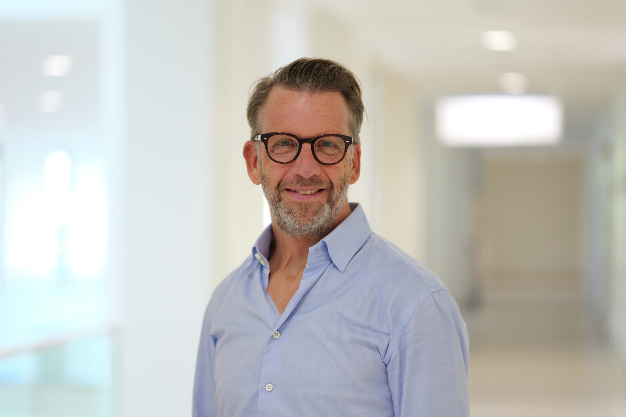 Dr. Hanke, ein freundlicher Mann mit Brille und kurzem Bart trägt ein hellblaues Hemd und steht in einem hell ausgeleuchteten Klinikflur. Er lächelt leicht und blickt direkt in die Kamera.