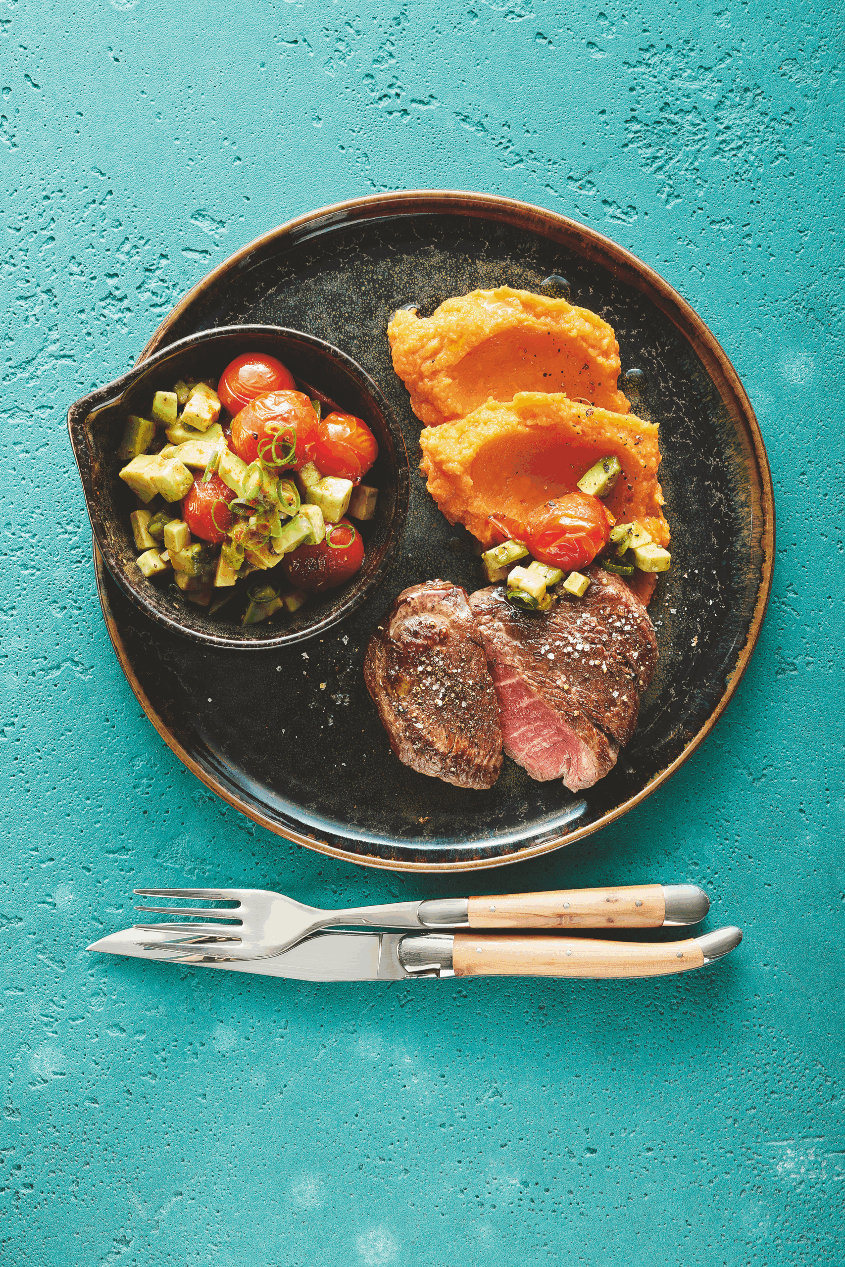 Medium gebratenes Rindersteak auf cremigem Süßkartoffelpüree mit Kräutergarnitur, serviert auf einem dunklen Teller.