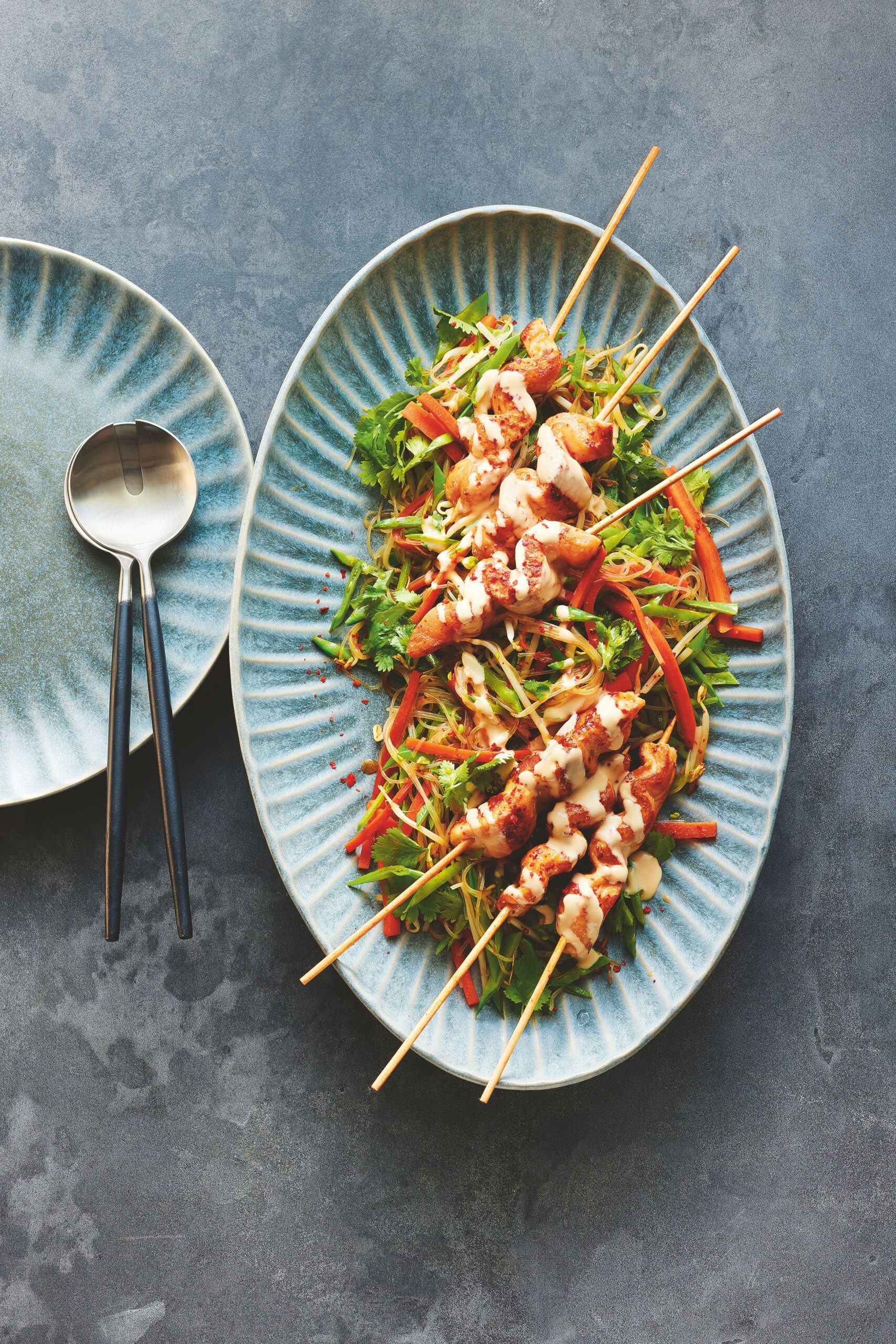 Asiatisches Fisch-Saté auf Glasnudelsalat mit Koriander, Paprika und cremigem Dressing auf blauem Keramikteller angerichtet.