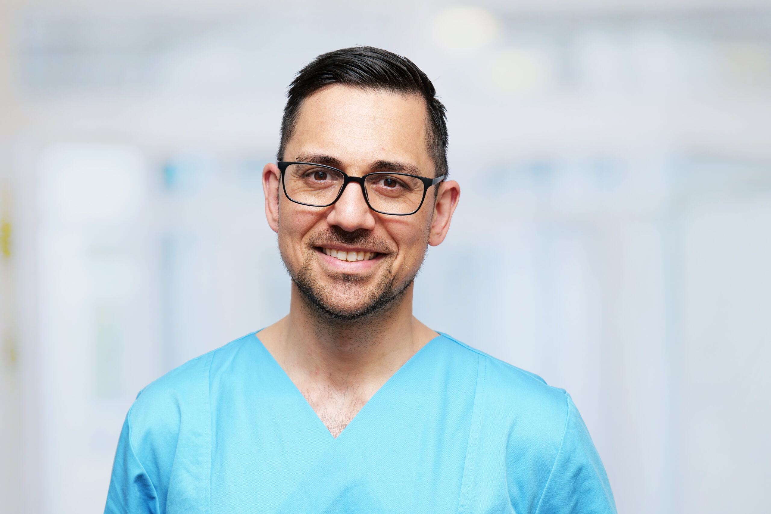 Dr. Ammar Moubayed, lächelnder Arzt in blauer OP-Kleidung, mit Brille, vor einem hellen Klinik-Hintergrund