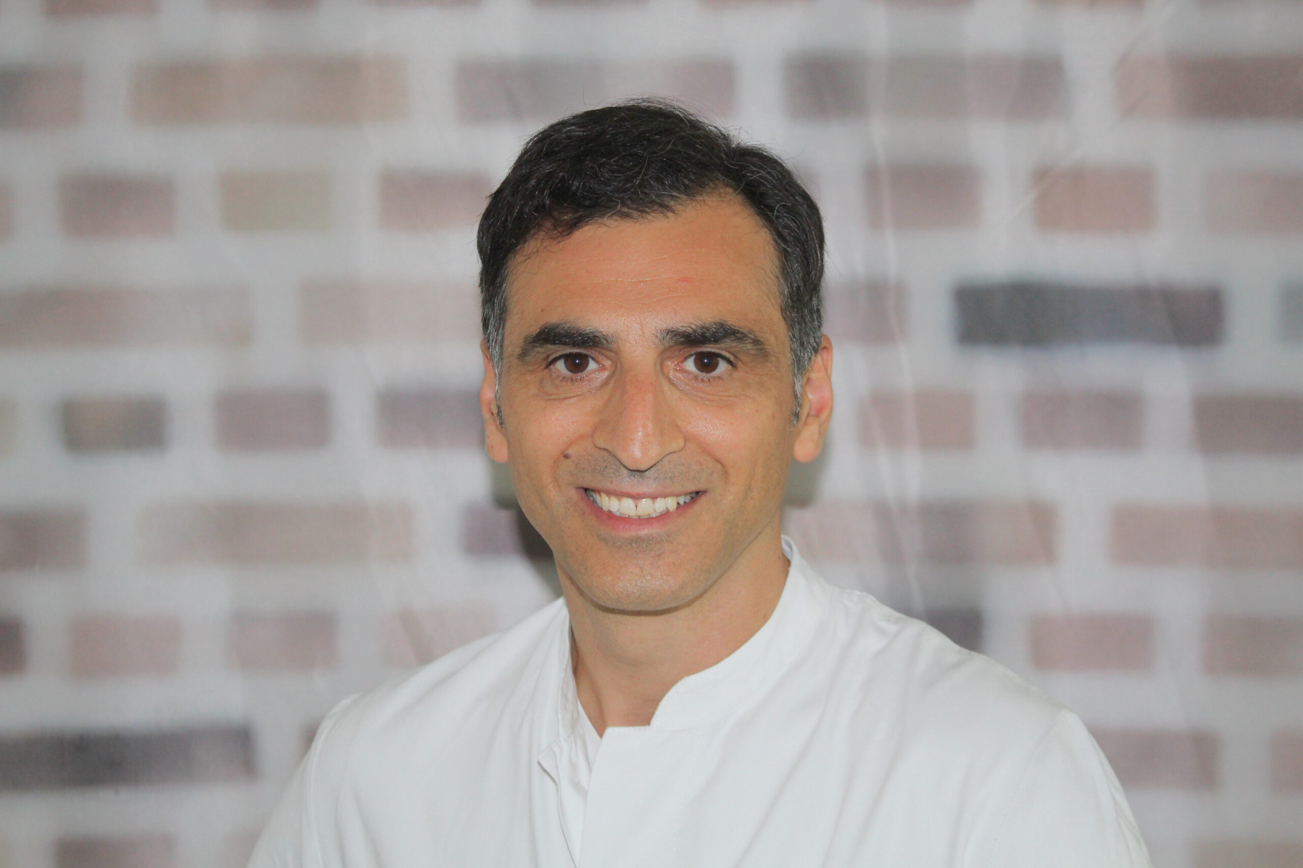 Porträt von Dr. Halil Dag in einem weißen Kittel vor einer Backsteinwand