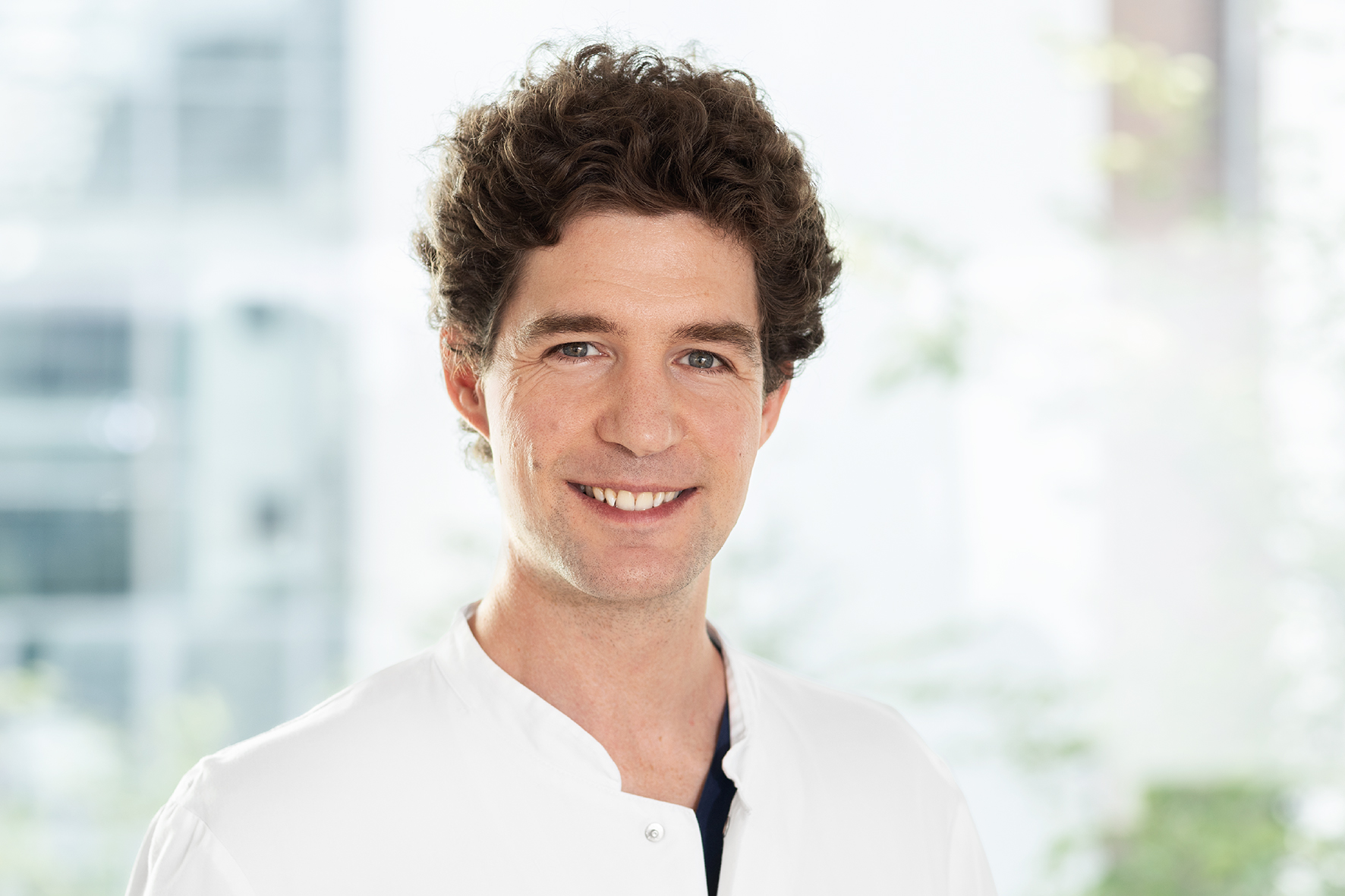 Junger Arzt im weißen Kittel, lächelnd vor hellem Hintergrund.