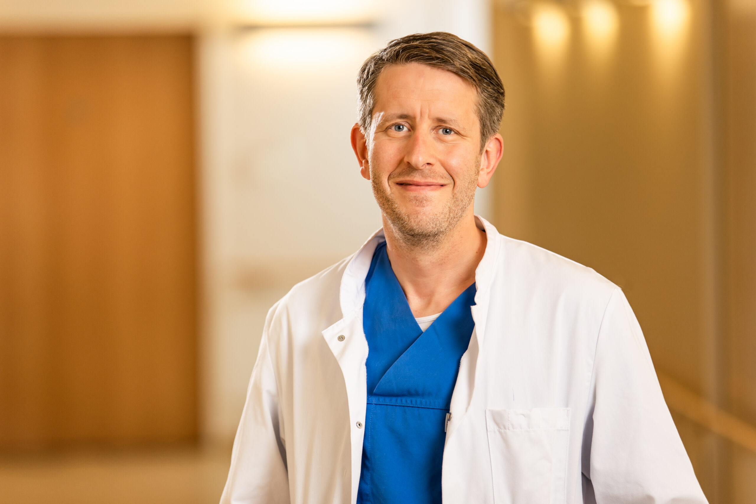 Arzt im weißen Kittel mit blauer OP-Kleidung, lächelnd im Krankenhausflur.