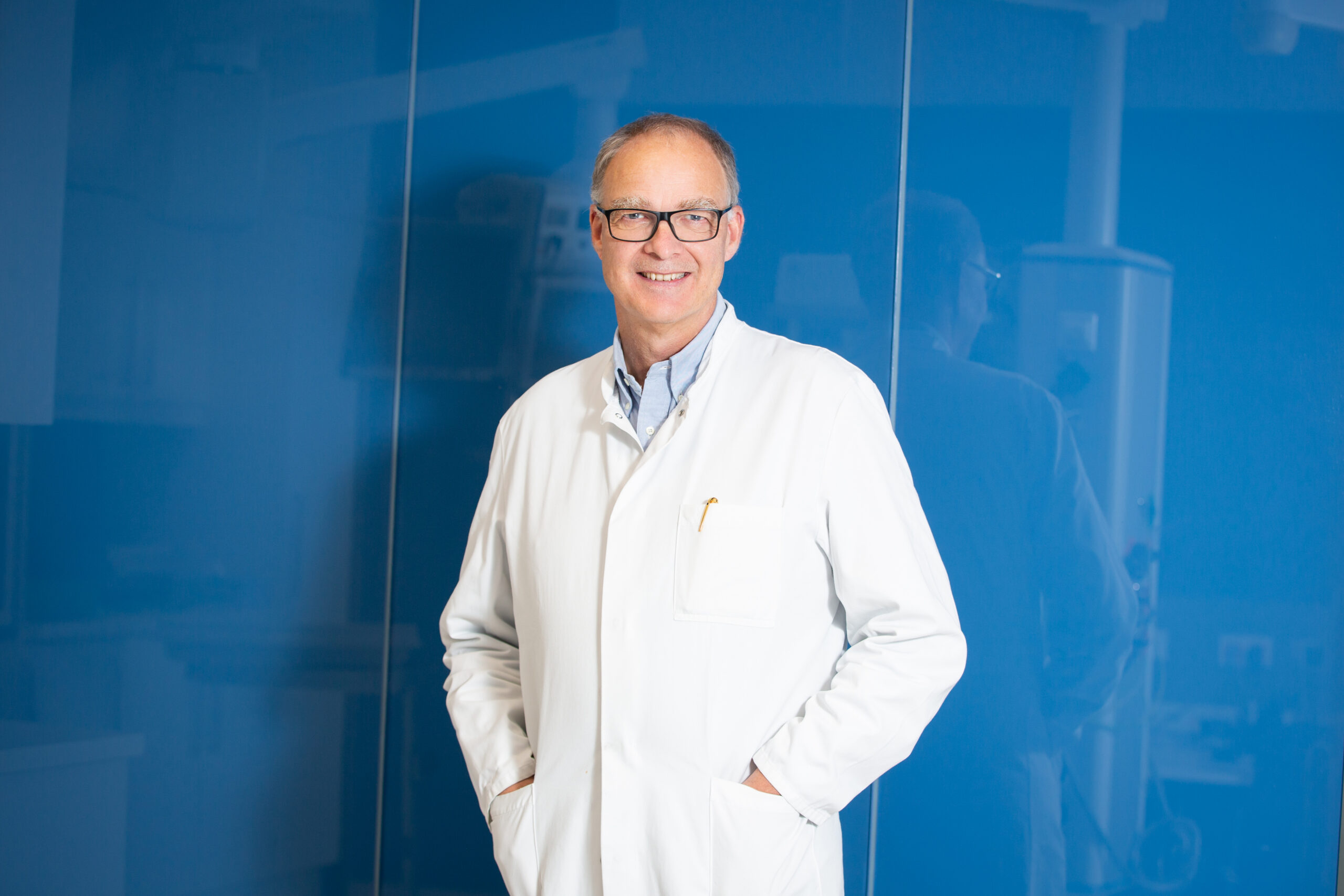 Lächelnder Arzt mit Brille im weißen Kittel vor blauer Glaswand, Hände in den Taschen.