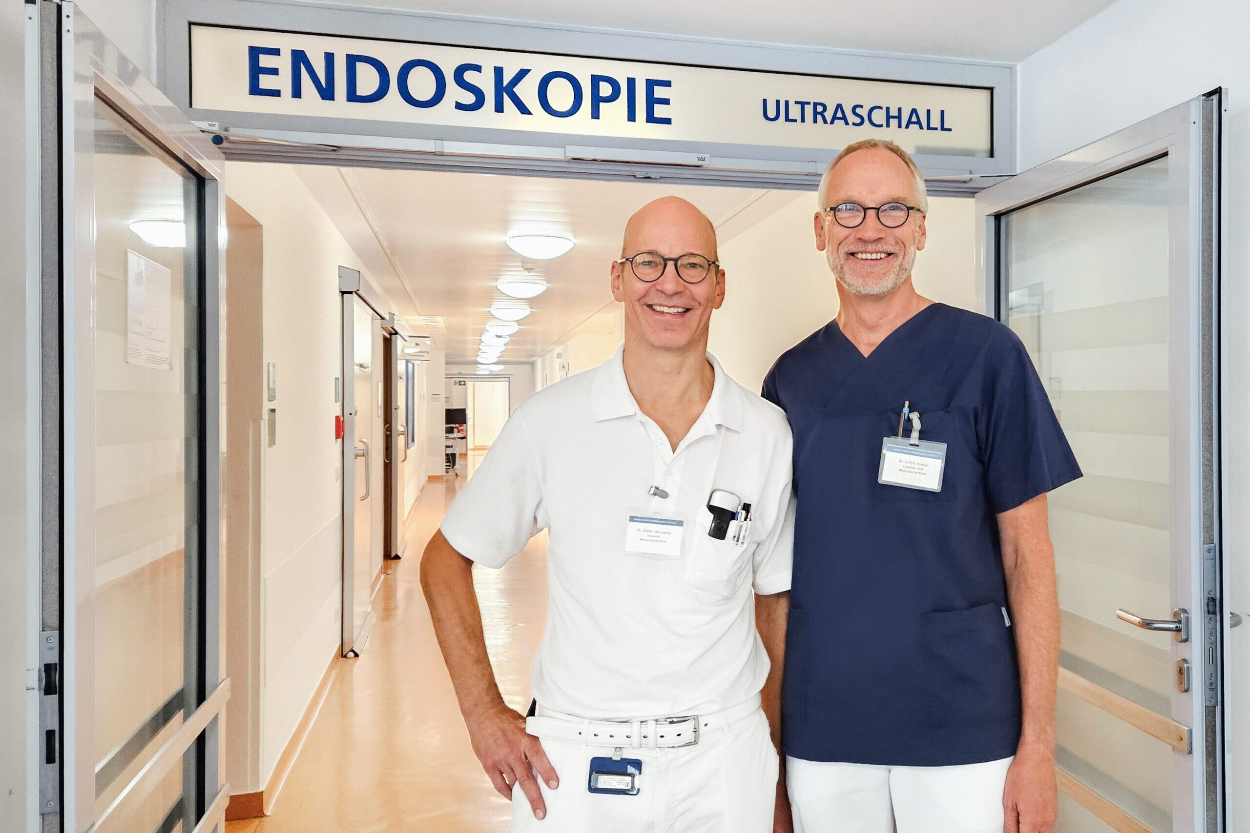 Zwei Männer in jeweils weißer und dunkelblauer Kleidung stehen in einem Krankenhausflur unter einem Schild mit der Aufschrift "Endoskopie".
