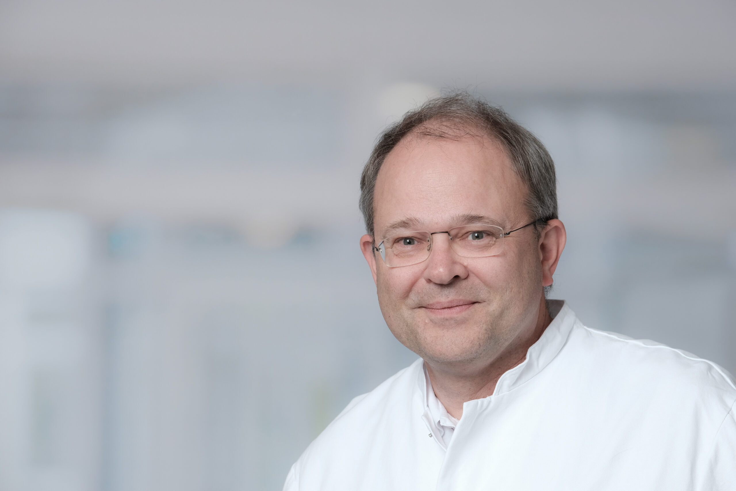 Porträt eines lächelnden Mannes (Prof. Dr. Guntram Lock) mittleren Alters mit Brille und kurzem Haar in weißem ärztlichem Kittel vor einem neutralen, unscharfen Hintergrund – vermutlich in einer medizinischen Einrichtung aufgenommen.