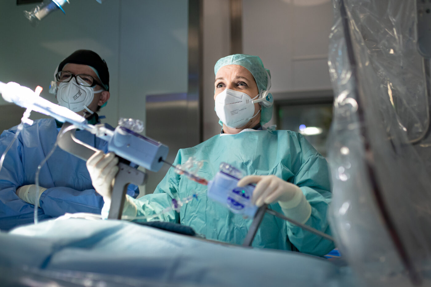 Zwei Personen in OP-Kleidung während eines medizinischen Eingriffs. Im Vordergrund eine Frau mit OP-Maske, Haube und Headset, bei der Einsetzung eines Mitralklappen-Clips.