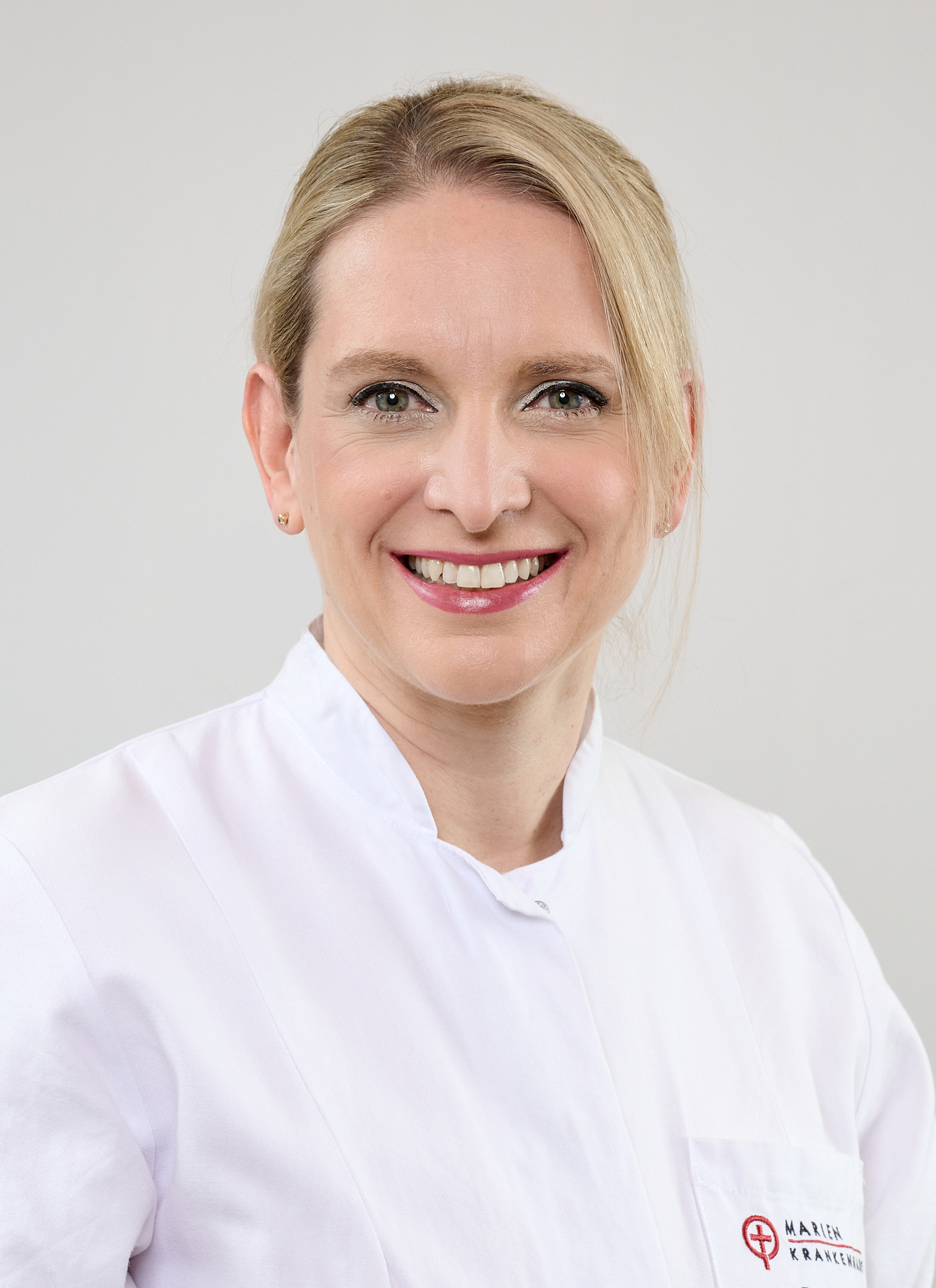 Porträt einer freundlich lächelnden Frau (Prof. Dr. Lubos) in weißem ärztlichem Kittel mit aufgesticktem Logo „Marienkrankenhaus“. Sie hat hellblonde, seitlich zurückgebundene Haare und trägt dezente Ohrringe. Heller, neutraler Hintergrund.