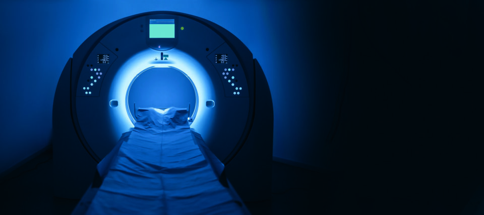 Ein leerer, in Blau beleuchteter Computertomograph (CT) in einem abgedunkelten Raum. Die Untersuchungsliege ist in die Öffnung des Geräts eingefahren.