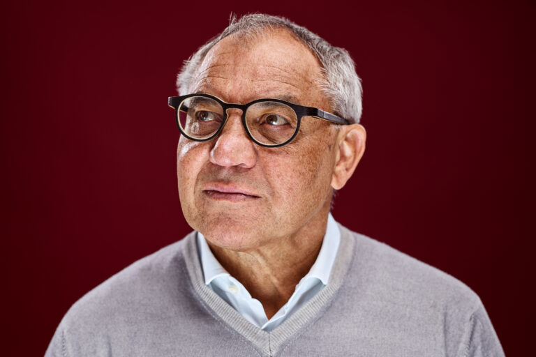 Felix Magath schaut in die obere linke Ecke. Er trägt einen grauen Pulunder, aus dem ein hellblauer Hemdkragen herausragt. Der Hintergrund ist weinrot.