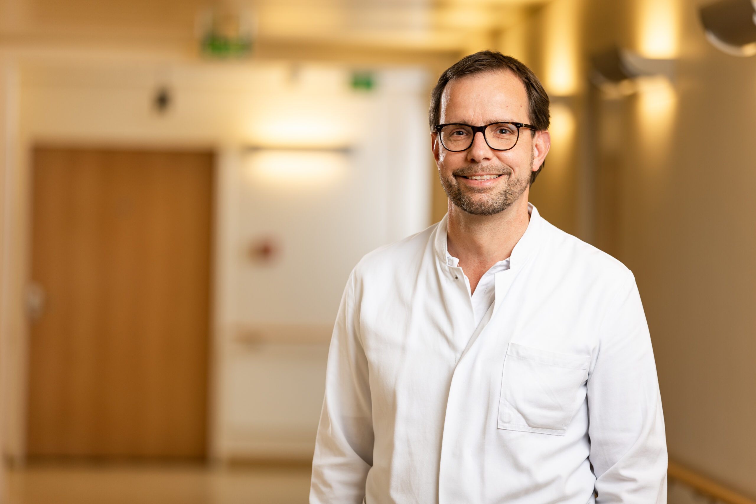 Der Arzt, Dr. Frank Trostdorf, mit kurzen dunklen Haaren, leichtem Bart und schwarzer Brille steht in einem warm beleuchteten Krankenhausflur und blickt freundlich in die Kamera. Er tr&auml;gt einen wei&szlig;en Arztkittel &uuml;ber einem hellen Hemd.