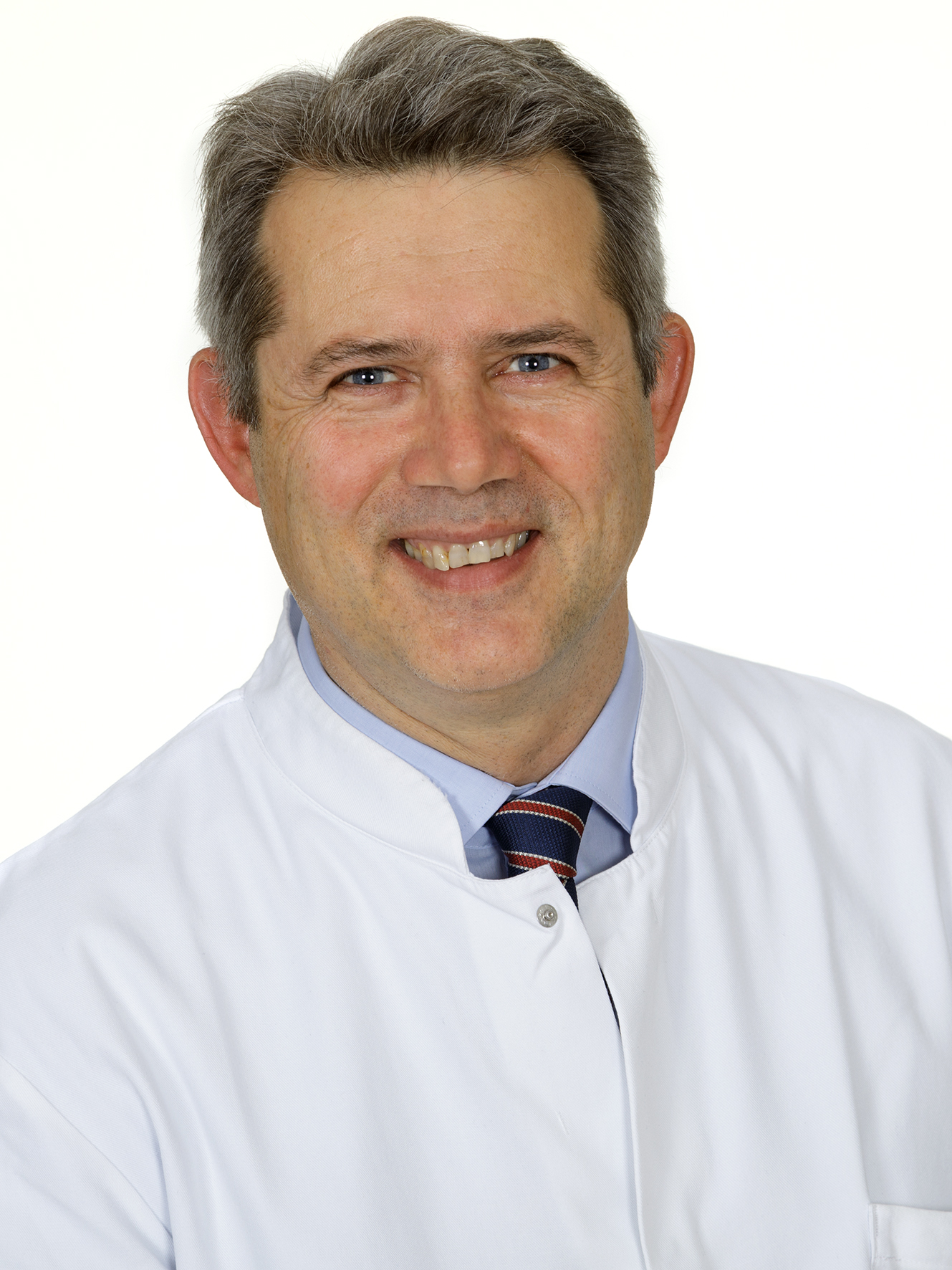 Porträt des Arztes Dr. Florian Lienau mit kurzen braunen Haaren und freundlichem Lächeln vor hellem Hintergrund. Er trägt einen weißen Arztkittel über einem hellblauen Hemd mit gestreifter Krawatte und blickt direkt in die Kamera.