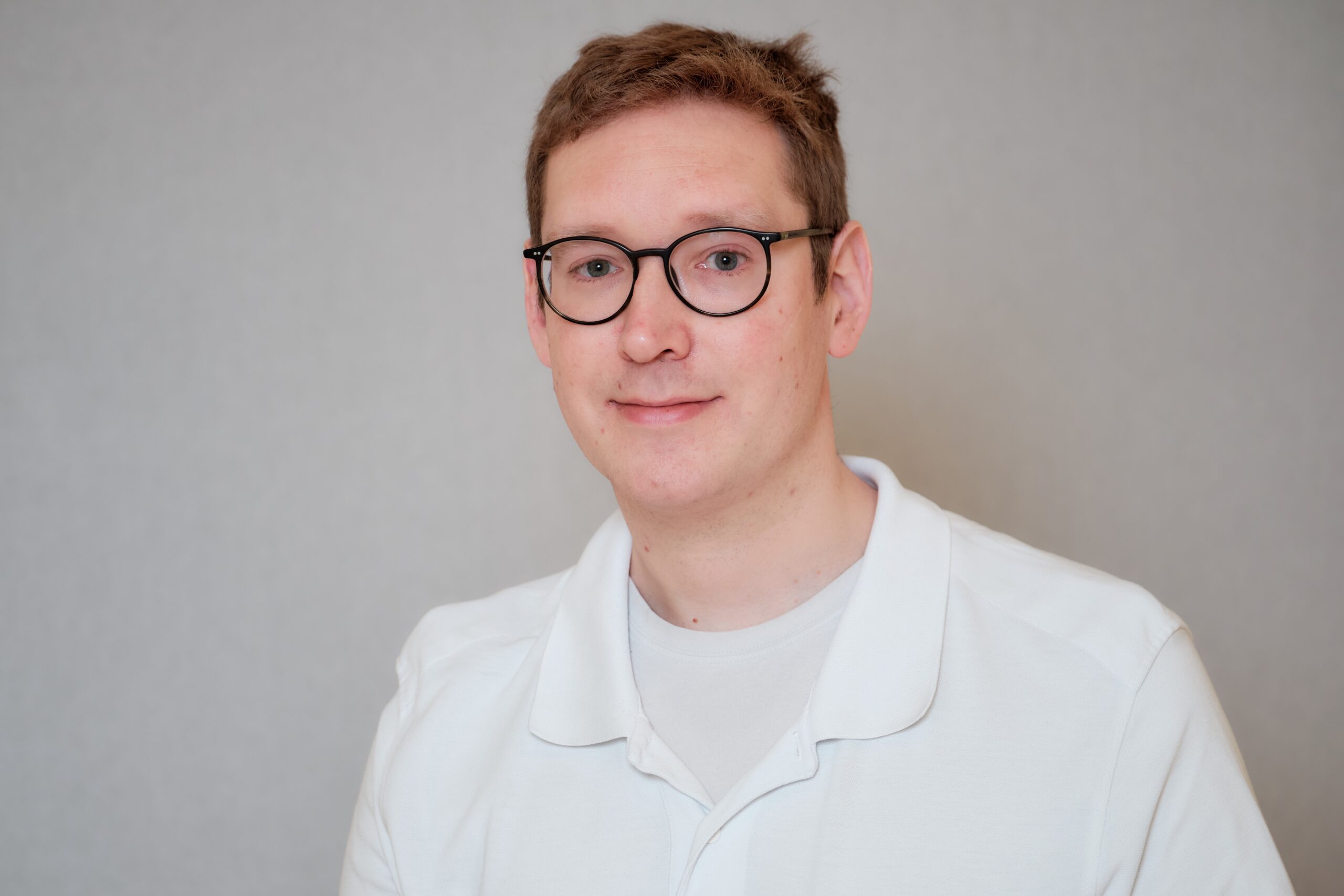 Porträt von Dr. Peer Spyra mit kurzen hellbraunen Haaren und runder schwarzer Brille vor einem neutral grauen Hintergrund.