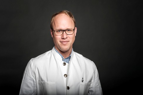 Porträt von Prof. Dr. Felix Hilpert im weißen Arztkittel vor dunklem Hintergrund.