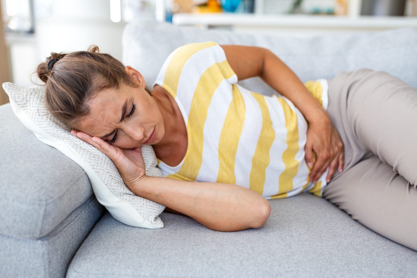Frau liegt mit geschlossenen Augen auf einem Sofa und hält sich wegen Bauchschmerzen den Bauch.